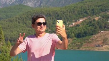 Man taking selfie on mobile phone of mountains lake background. Traveler male having video chat on cellphone on the blue lake outdoors travel adventure vacation.