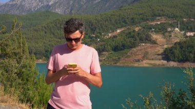 Man texting online on mobile phone of mountains lake background. Traveler male chat messages on cellphone on the blue lake outdoors travel adventure vacation. Using smartphone