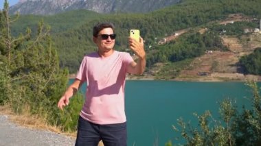 Man taking selfie on mobile phone of mountains lake background. Traveler male having video chat on cellphone on the blue lake outdoors travel adventure vacation.