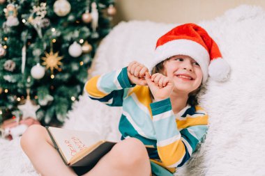 Kırmızı Noel Baba şapkalı gülen çocuk kanepede oturup sevgili Noel Baba 'ya mektup yazıyor. Çocuk dilek listesi. Noel hediyelerinin rüyaları. Mutlu Noeller ve mutlu yıllar.