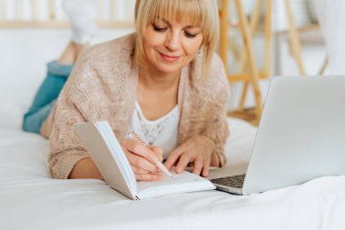 Çevrimiçi eğitim ve çalışma mesafesi. Yetişkin bir kadın yatağa uzanıyor, deftere yazıyor. Ev ofisinde dizüstü bilgisayarı olan serbest çalışan biri. Bilgisayar ve online alışveriş kullanarak..