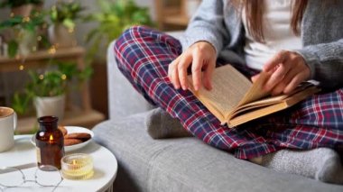 Sıcak kadın bacakları örülmüş sıcak çorap ve kareli pijama ile kitap okuyup sayfaları karıştırıyor, kupa içinde kahve içiyor, evdeki kanepede oturuyor. Mum, dekor ve kapalı alan bitkileriyle sonbahar titreşimleri