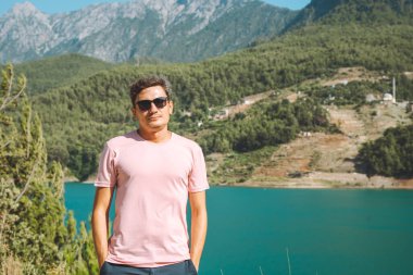 Arka planda, dağların yakınında duran, güneş gözlüklü gülümseyen adamın portresi. Mavi gölde seyahat eden genç bir erkek macera tatili yapıyor..