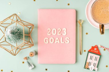 Christmas pink notepad with 2023 goals letters text. Flat lay on green mint table background with planner, cup of coffee, candle, Christmas decoration, notebook and stationery. Top view Desktop.