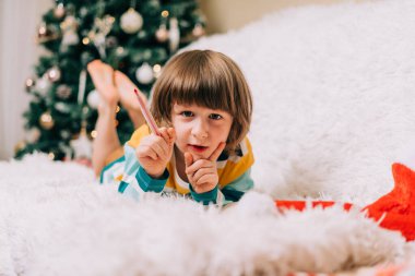 Gülümseyen çocuk kanepeye uzanıp Noel ağacının yanındaki Noel Baba 'ya mektup yazıyor. Çocuk dilek listesi. Noel hediyelerinin rüyaları. Mutlu Noeller ve mutlu yıllar.
