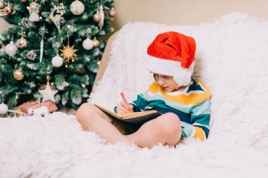 Kırmızı Noel Baba şapkalı gülümseyen çocuk kanepede oturup sevgili Noel Baba 'ya mektup yazıyor. Çocuk dilek listesi. Noel hediyelerinin rüyaları. Mutlu Noeller ve mutlu yıllar.
