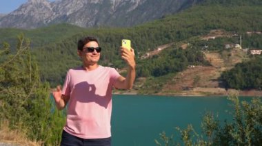 Man taking selfie on mobile phone of mountains lake background. Traveler male having video chat on cellphone on the blue lake outdoors travel adventure vacation.