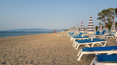 Mavi ve beyaz şemsiye ve plajda mavi deniz ve mavi gökyüzünün önünde plaj sandalyeleri. Calabria, Simeri Mare, İtalya.