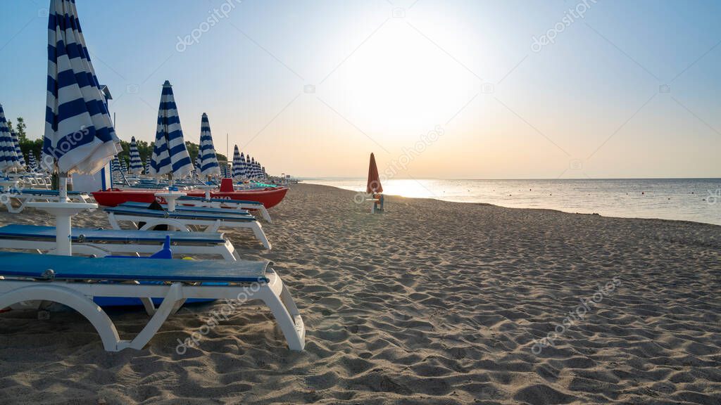 Blue and white umbrella and beach chairs at the beach in front of adriatic sea at sunrise. Calabria, Simeri Mare, Italy.