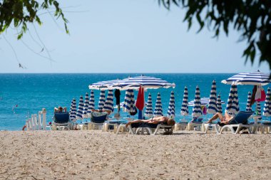 Mavi ve beyaz şemsiye ve kumsalda mavi deniz ve mavi gökyüzünün önünde plaj sandalyesi. Calabria, Simeri Mare, İtalya.