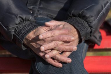 Yaşlı bir adam bankta oturur ve ellerini dizlerinin üstüne koyar.