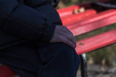 Yaşlı bir adam bankta oturur ve ellerini dizlerinin üstüne koyar.