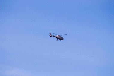 Siyah bir helikopter ormanın üzerinde mavi gökyüzünde uçuyor.