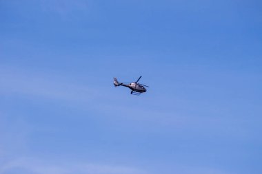 Siyah bir helikopter ormanın üzerinde mavi gökyüzünde uçuyor.