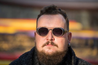 Siyah gözlüklü, sakallı ve bıyıklı genç bir adam. Adam bir şeyler düşünmüş.