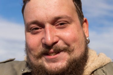 Kulağında bıyığı, sakalı ve orta parmağı olan genç bir adam gülümsüyor ve mutlu.