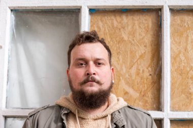 Bıyıklı ve sakallı genç bir adam kafası karışmış ve üzgün. Ahşap bir pencerenin arkasındaki adam