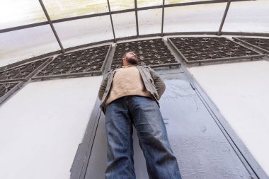 Sakallı genç bir adam yarı dairesel bir çatı altında duruyor.