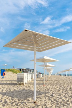 Deniz kıyısında güneşten ahşap şemsiyeler. Plaj sezonu kapanıyor. Plajda tatilciler için beyaz tasarımlar. Kumlu sahilde tatilciler için güneşten korunacak barınaklar var..