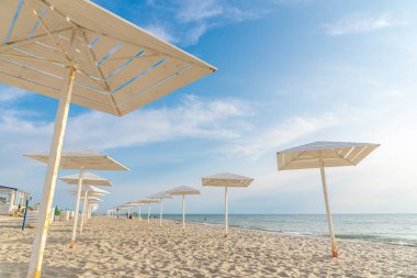 Deniz kıyısında güneşten ahşap şemsiyeler. Plaj sezonu kapanıyor. Plajda tatilciler için beyaz tasarımlar. Kumlu sahilde tatilciler için güneşten korunacak barınaklar var..
