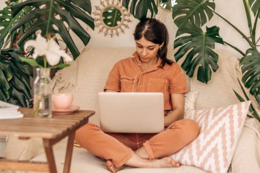 Koyu renk saçlı, dizüstü bilgisayar kullanan genç bir kadın. Arka planda iç mekan bitkileri. İş, online alışveriş, e-öğrenme, kentsel orman kavramı..