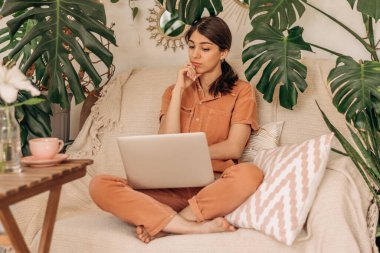 Genç düşünceli melez kadın evde koltukta otururken dizüstü bilgisayar kullanıyor. Arka planda kapalı bitkiler. İş, online alışveriş, e-öğrenme, kentsel orman konsepti..