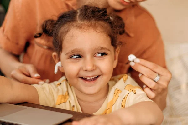 Close-up of a small laughing mixed-race girl sitting with her mother in front of a laptop.Mom inserts wireless headphones into the girl\'s ears.Working at home mom.Gadgets and children concept.