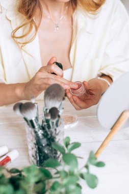 Beyaz bir masada makyaj fırçaları, ayna ve dekoratif kozmetik ürünleri. Arka plandaki bulanık kadın elleri tüpü aç. Güzellik kavramı..