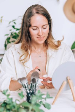 Güzel genç bir kadın evde aynanın önünde makyaj yapıyor. Evde elf bakımı. Güzellik konsepti..