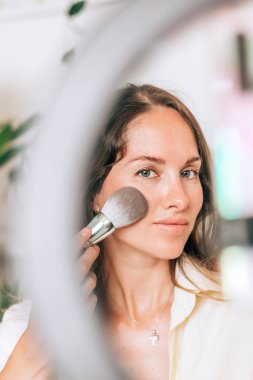 Genç bir kadın güzellik blogcusu, sosyal medyada makyaj ve kozmetik ürünlerini incelemek için canlı yayın yapıyor, video blogu için güzellik ürünlerini gözden geçiriyor. Nüfuzlu kişi tripod ve halka lamba üzerinde akıllı telefon kullanıyor.