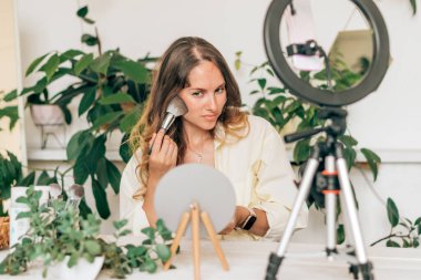 Genç bir kadın güzellik blogcusu, sosyal medyada makyaj ve kozmetik ürünlerini incelemek için canlı yayın yapıyor, video blogu için güzellik ürünlerini gözden geçiriyor. Nüfuzlu kişi tripod ve halka lamba üzerinde akıllı telefon kullanıyor.