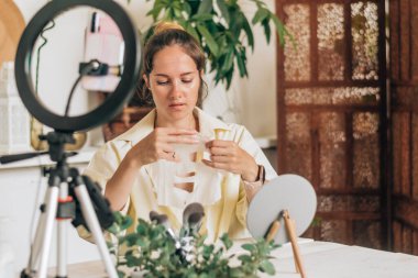 Genç kadın güzellik blogcusu, makyajını ve kozmetik ürünlerini sosyal medyada incelemek için canlı yayın yapıyor, yüzüne kumaş kozmetik maske takıyor. Etki cihazı tripod ve halka lamba üzerinde akıllı telefon kullanıyor.