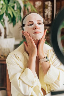 Genç kadın güzellik blogcusu, makyajını ve kozmetik ürünlerini sosyal medyada incelemek için canlı yayın yapıyor, yüzüne kumaş kozmetik maske takıyor. Etki cihazı tripod ve halka lamba üzerinde akıllı telefon kullanıyor.