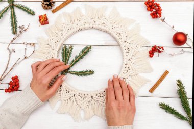 Kadın makrome tarzında bir Noel çelengini köknar dalları ve yaban mersini ile süslüyor, el yapımı eko-stil dekor, eller yakın çekim, üst manzara. Noel, yeni yıl ve çevre dostu konsept..