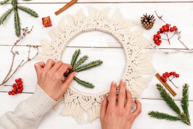 Kadın makrome tarzında bir Noel çelengini köknar dalları ve yaban mersini ile süslüyor, el yapımı eko-stil dekor, eller yakın çekim, üst manzara. Noel, yeni yıl ve çevre dostu konsept..