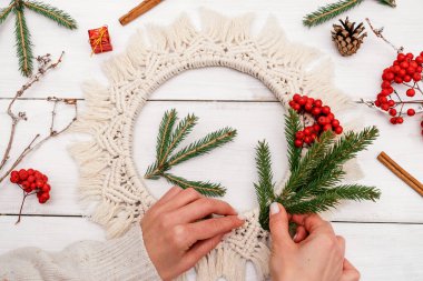 Kadın makrome tarzında bir Noel çelengini köknar dalları ve yaban mersini ile süslüyor, el yapımı eko-stil dekor, eller yakın çekim, üst manzara. Noel, yeni yıl ve çevre dostu konsept..