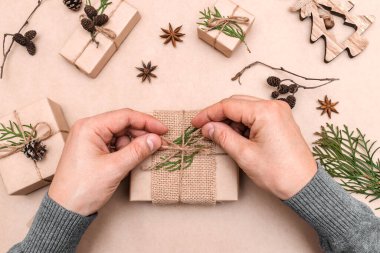 Hediyelerin eko-stil dekorasyonu, erkek eli yakın çekim, üst görünüm. Hediye kutuları el sanatları kağıdına sarılmış, thuja yaprakları ve koni ile süslenmiş. Noel, yeni yıl ve çevre dostu kavram..
