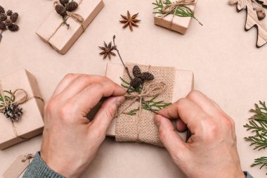Hediyelerin eko-stil dekorasyonu, erkek eli yakın çekim, üst görünüm. Hediye kutuları el sanatları kağıdına sarılmış, thuja yaprakları ve koni ile süslenmiş. Noel, yeni yıl ve çevre dostu kavram..