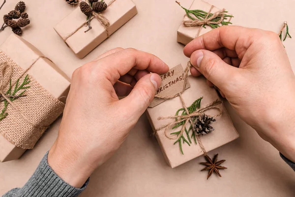 Hediyelerin eko-stil dekorasyonu, erkek eli yakın çekim, üst görünüm. Hediye kutuları el sanatları kağıdına sarılmış, thuja yaprakları ve koni ile süslenmiş. Noel, yeni yıl ve çevre dostu kavram..