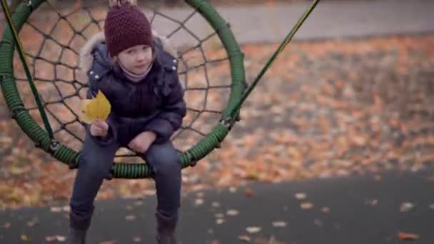 Unscharfes 4-5 jähriges Mädchen in Jacke und Hut reitet auf einer Schaukel auf dem Spielplatz — Stockvideo