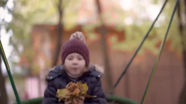 Chica borrosa de 4-5 años en una chaqueta y un sombrero cabalga en un columpio en el patio de recreo — Vídeo de stock