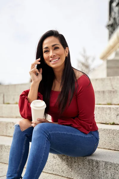 Beautiful Latina woman sitting on the street stairs on a spring day and having a mobile conversation on the smartphone while holding a coffee in her hand. — Stock Photo, Image