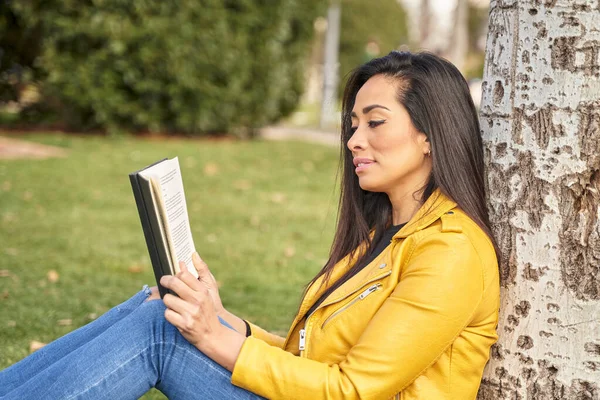 Donkerharige vrouw zittend in het gras, leunend tegen een boom, een boek lezend op een lentedag in een park. — Stockfoto