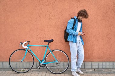 Genç, kıvırcık saçlı, kulaklıklı, dışarıda duvarda duran bisikletinin yanında cep telefonu kullanan bir öğrenci.