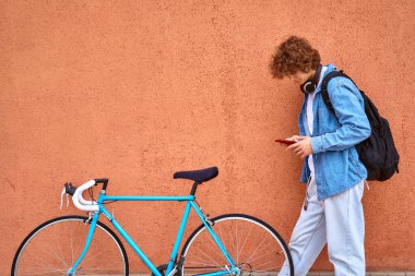 Genç, kıvırcık saçlı, kulaklıklı, dışarıda duvarda duran bisikletinin yanında cep telefonu kullanan bir öğrenci.