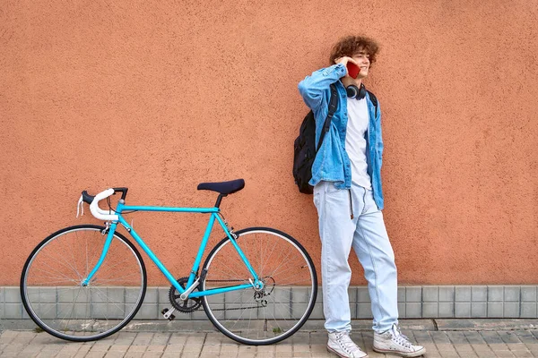 Tersenyum pemuda berambut keriting dengan headphone berdiri di dinding luar berbicara pada ponsel. — Stok Foto