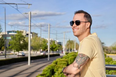 Güneş gözlüklü bir adam güneşli bir günde dışarıda dururken ufka bakıp gülümsüyor. Kentsel yaşam tarzı konsepti.