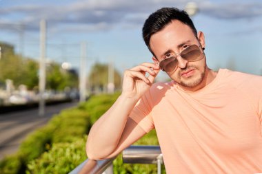 Ciddi bir genç adam güneşli bir günde şehir sokaklarında kameraya güneş gözlüğüyle bakıyor..