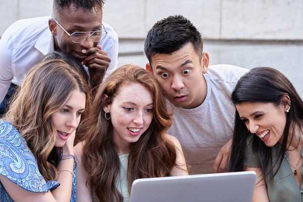 Group of multiracial people using a laptop computer together while sitting on stairs outdoors. Urban lifestyle, technology and friendship concept.