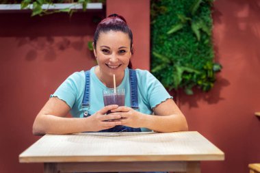 Young woman looking at the camera and smiling while drinking a fresh smoothie in a cafe. Healthy lifestyle and drinks concept.